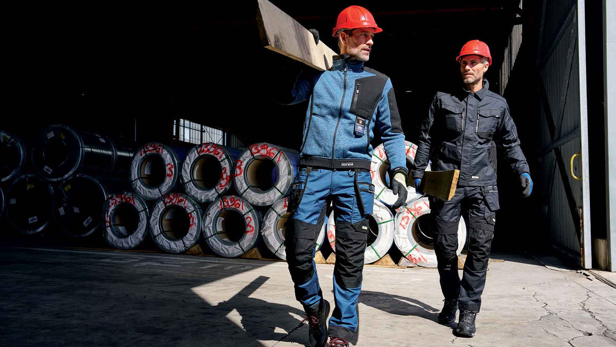 Two workers in Sir Safety Stretch Canvas workwear carrying timber through a steel warehouse