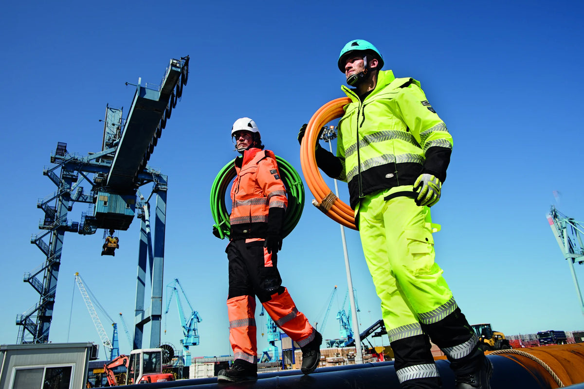 Two port workers in Sir Safety hi-vis jacket and trousers carrying cable reels at a harbour crane